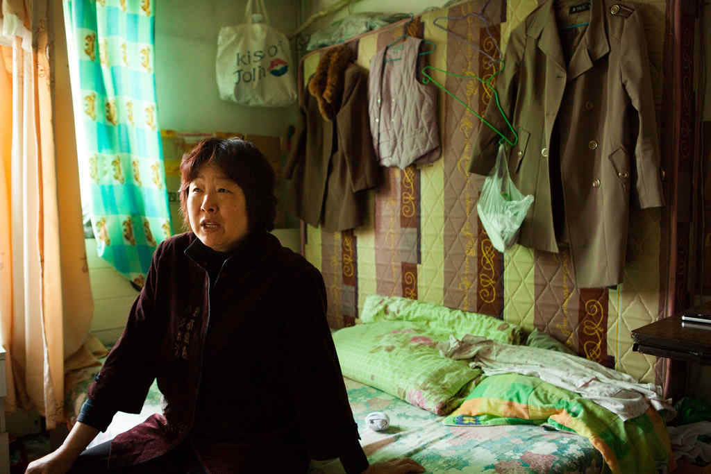 A middle aged Chinese woman sitting on a bed, with clothes hanging behind her