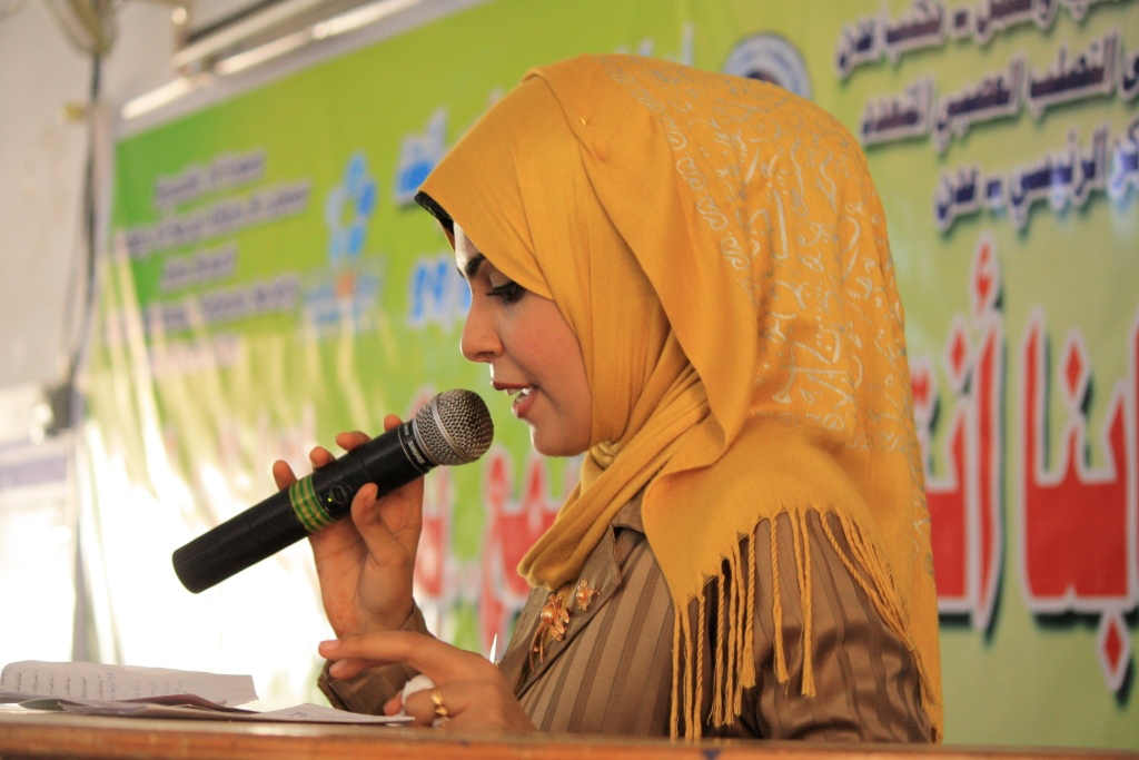 A woman in a yellow headscarf holds a microphone as she speaks to the room