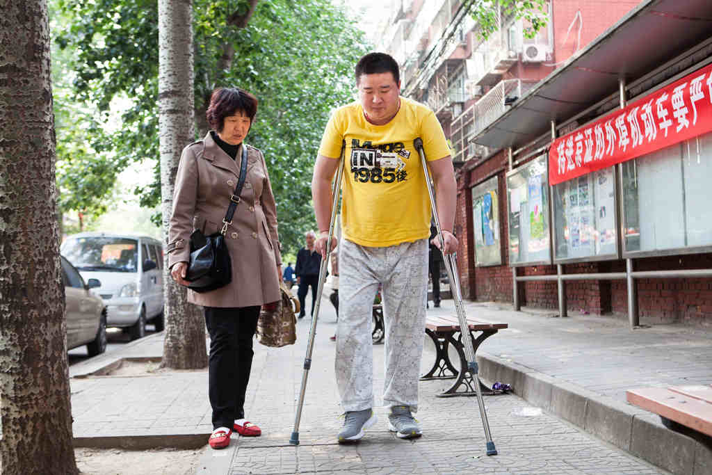 On a street in China a young man takes tentative steps using crutches. His mother looks on.