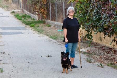 A woman stands on a small road with a dog. She is using a walking stick.