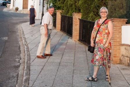 A woman stands smiling to the camera holding a wlaking stick. A man stands a few steps away looking at her and smiling.