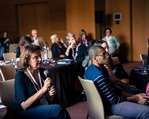 A woman sat in a chair speaks into a microphone while others sit listening in the background