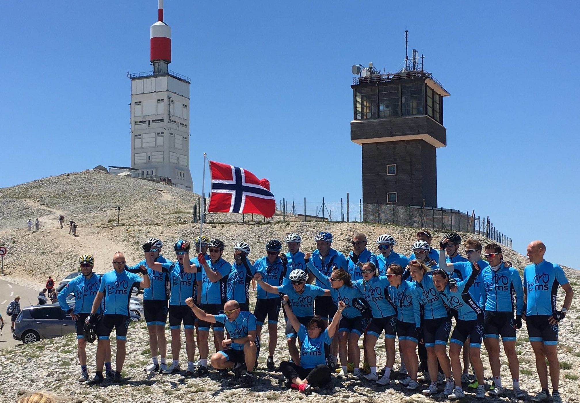 a dry sunny mountain top, a team of cyclists in blue tops pose for a picture in front of a Norwegian flag