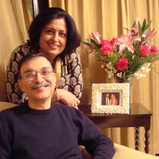 an Indian man and woman smile for the camera, behind them is a framed photo from their wedding day