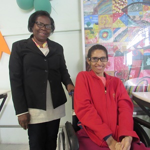 An older lady (Rita) stands with her hand on her adult daughter's wheelchair. They are both smiling at the camera.
