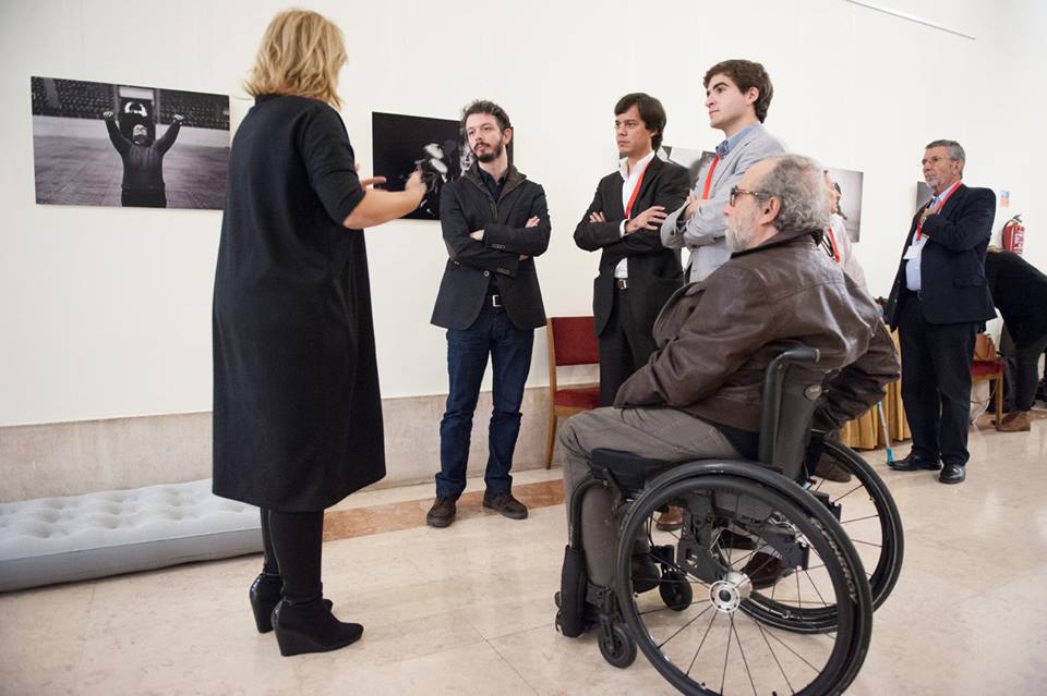 At the Portuguese Parliment a group of people are in a circle, most are standing, there is a man in a wheelchair in the foreground. There's an inflatable matress on the floor behind them
