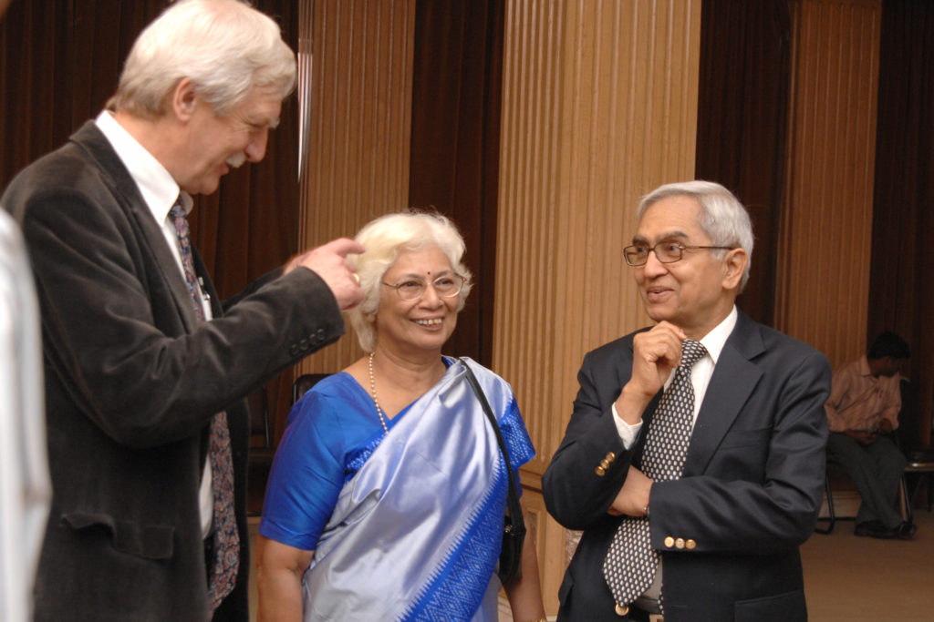 An Indian woman with white hair is smiling and talking with two men in suits